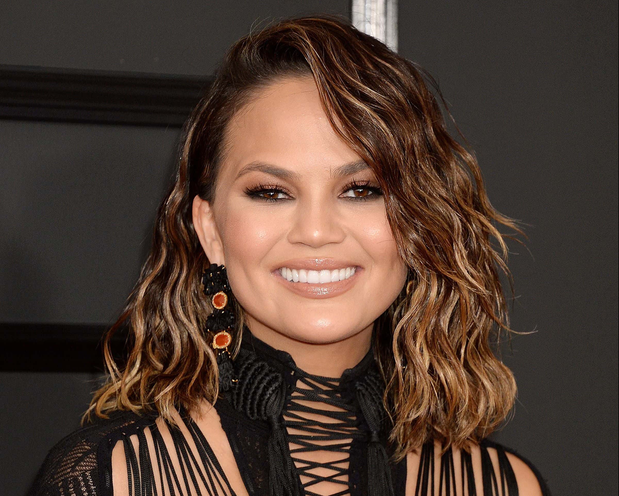 A modelo Chrissy Teigen posa sorrindo com cabelo curto ondulado castanho e roupa preta