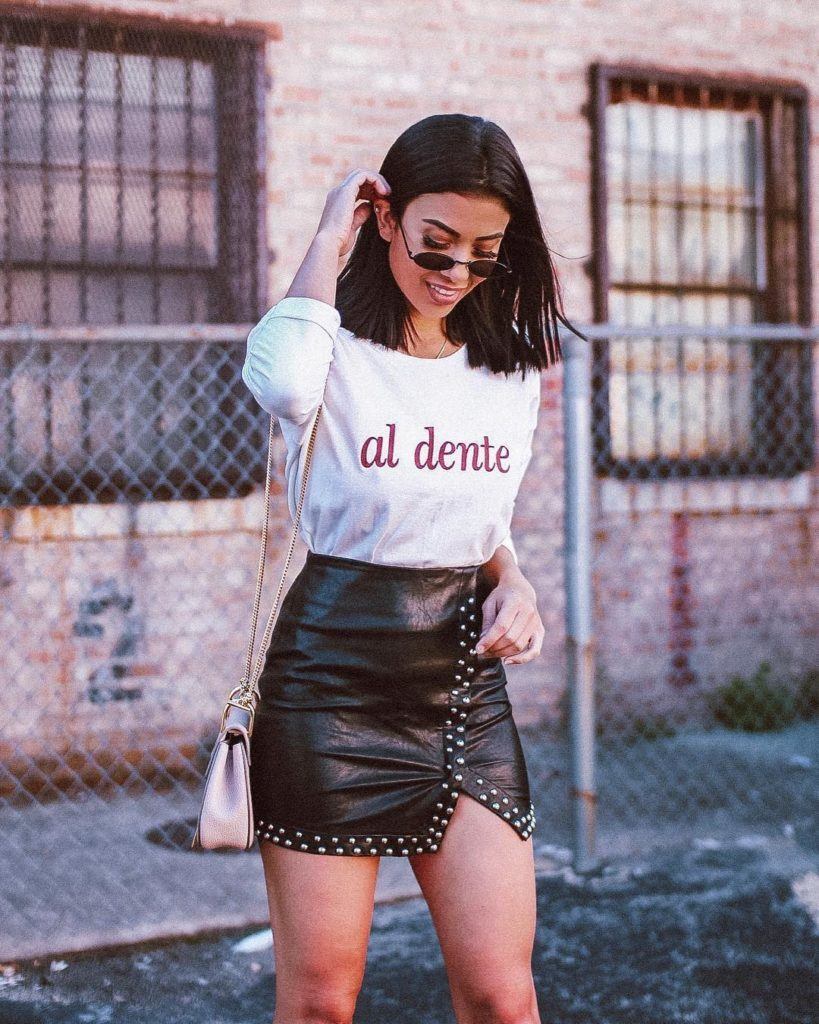 Mulher com cabelo castanho médio com óculos escuros pequenos, camiseta brança e minissaia de couro