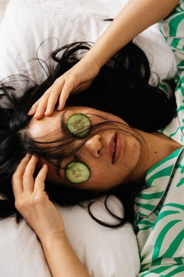 mulher deitada com o cabelo solto na cama e pepino nos olhos usando camisa estampada