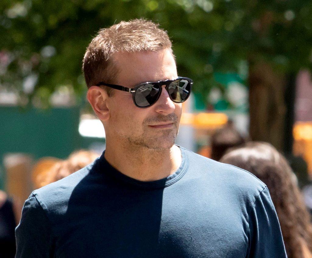Bradley Cooper caminha ao ar livre em dia de sol. Ele usa camiseta azul marinho e óculos de sol