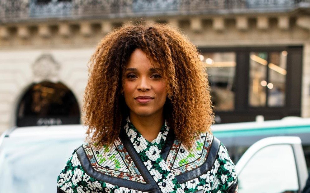 Modelo sorri para a foto. Ela tem cabelo cacheados soltos e usa uma blusa colorida, tipo quimono