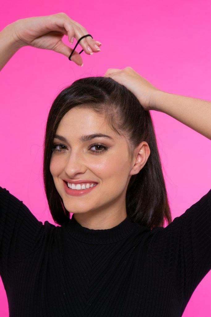 modelo de cabelo preto, liso e longo em fundo rosa fazendo um rabo de cavalo.