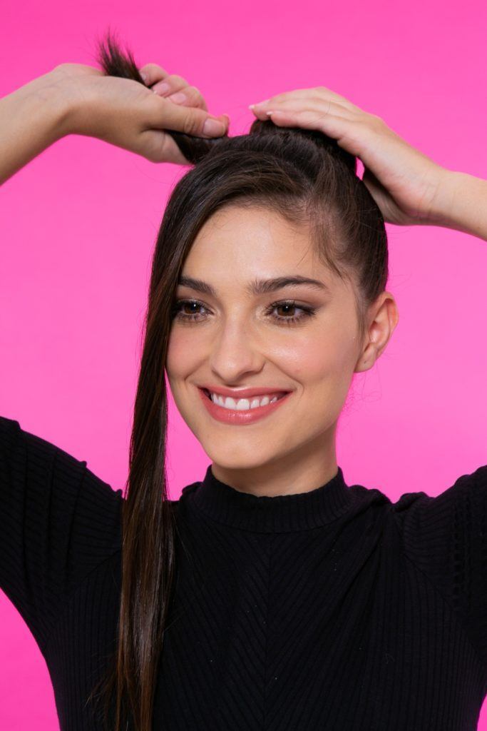 modelo de cabelo preto, liso e longo em fundo rosa fazendo um coque com uma mecha frontal solta