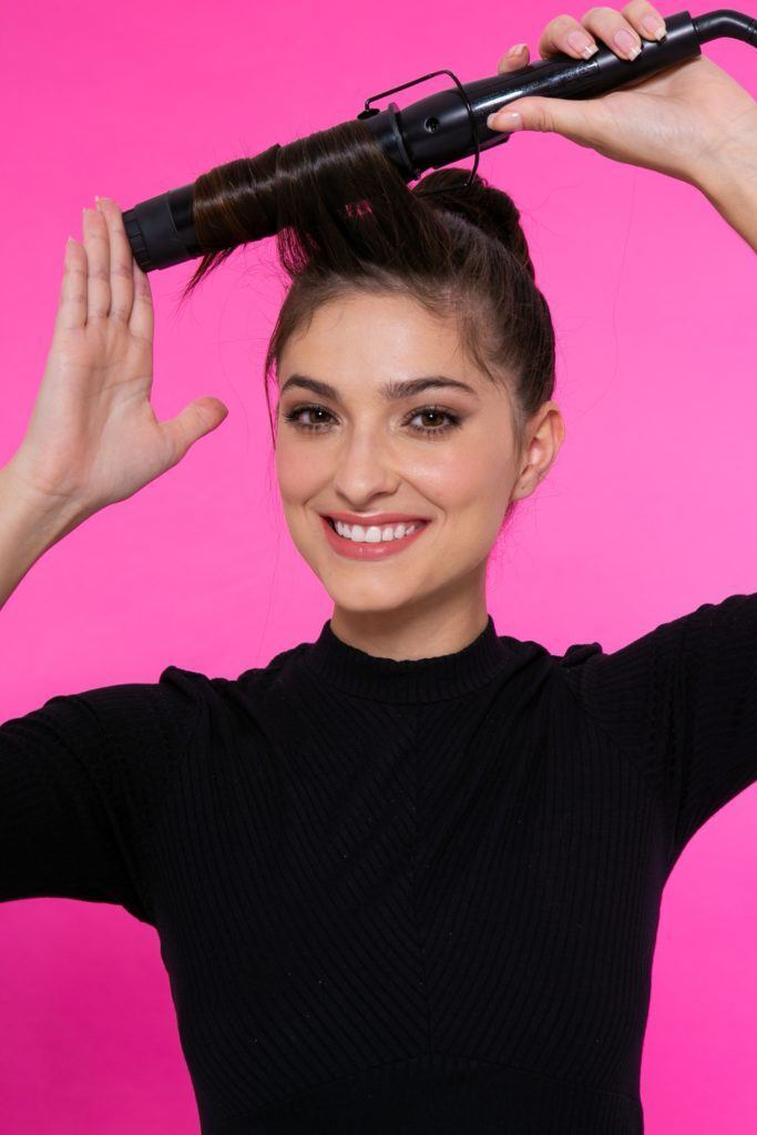 modelo de cabelo preto, liso e longo em fundo rosa com um coque, passando modelador de cachos em uma mecha frontal