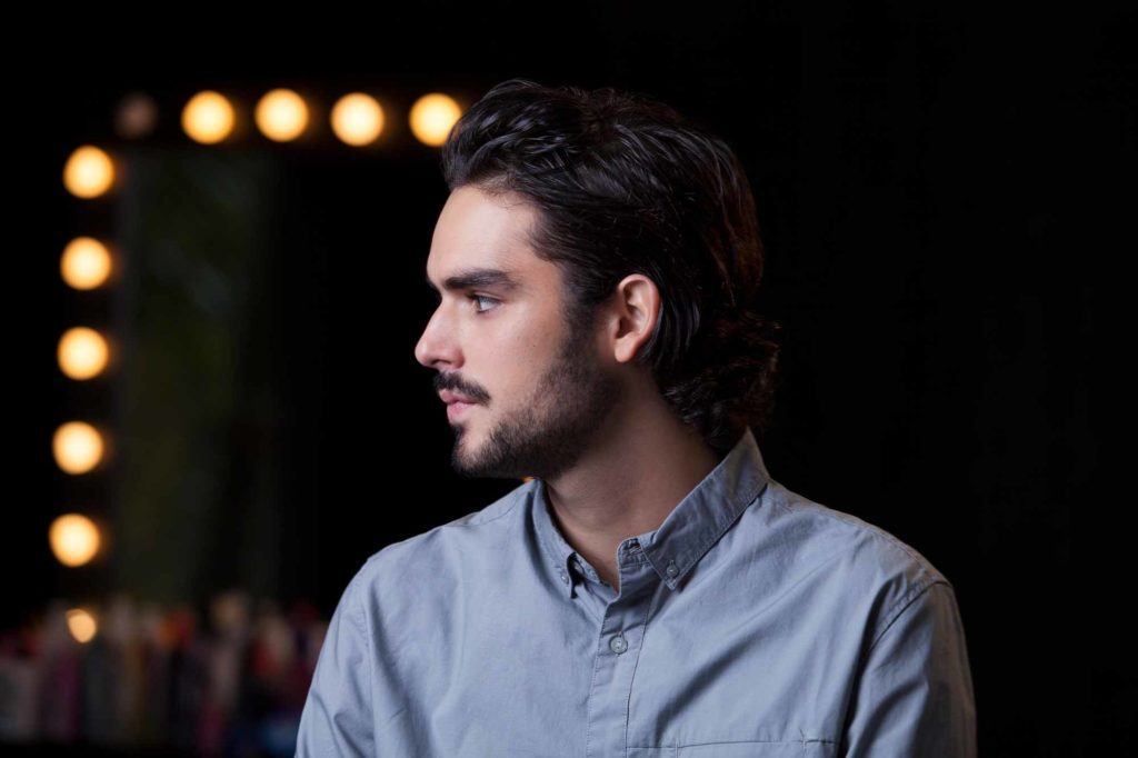 Homem usa slicked back hair para cabelo cacheado e camisa azul, contra o fundo preto com luzinhas amarelas