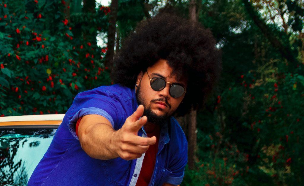 Homem negro de cabelo black power e usando óculos, ao ar livre. Ele está encostado no capô de um carro e há muitas árvores ao fundo