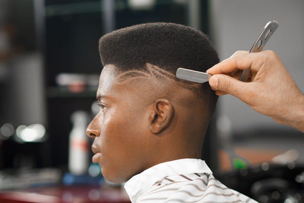 Homem negro no salão de barbeiro. Ele está sentado na cadeira, enquanto o profissional faz desenhos com navalha em seu cabelo