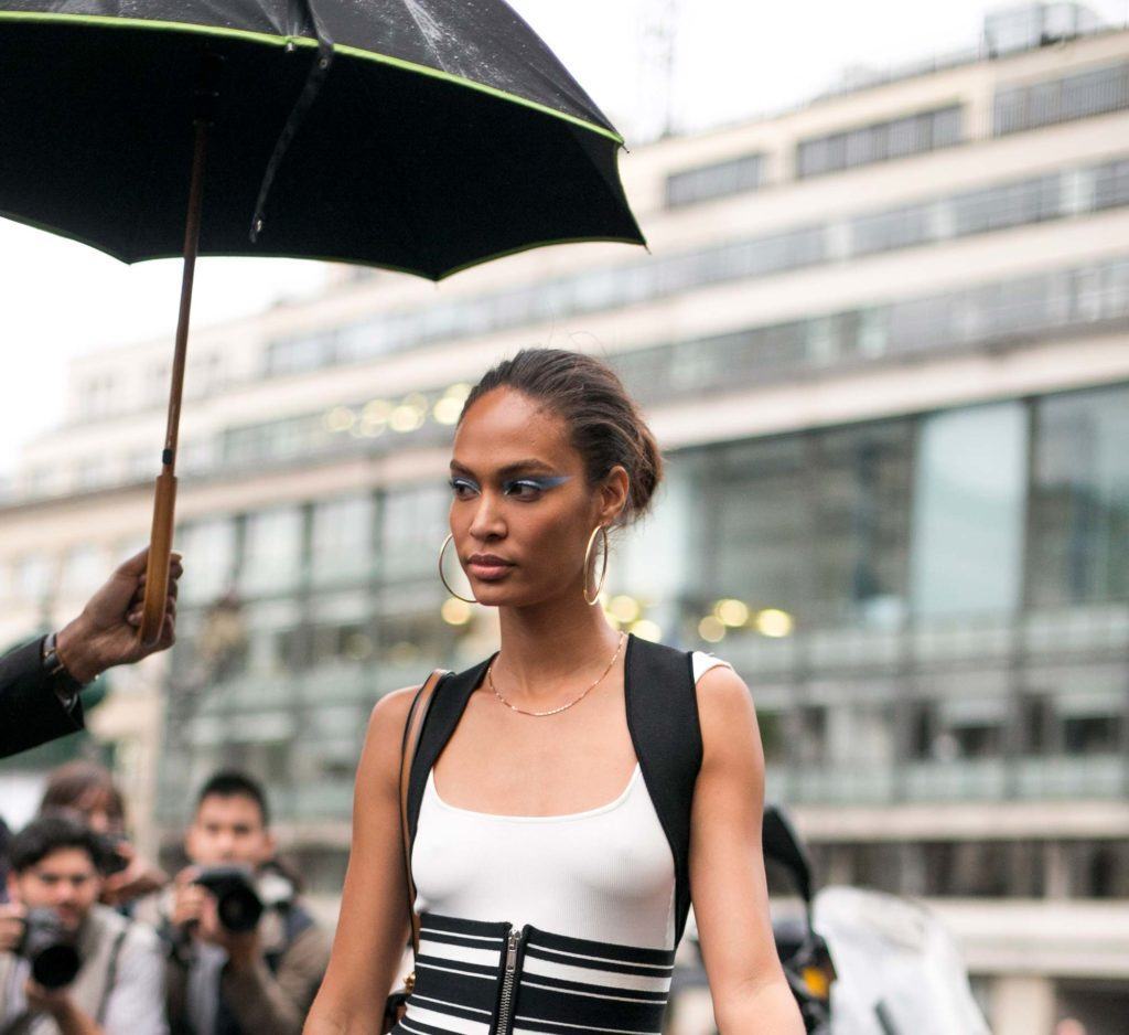 Mulher morena usa cabelo num coque, veste camiseta branca e tem alguém estendendo um guarda-chuva sobre si