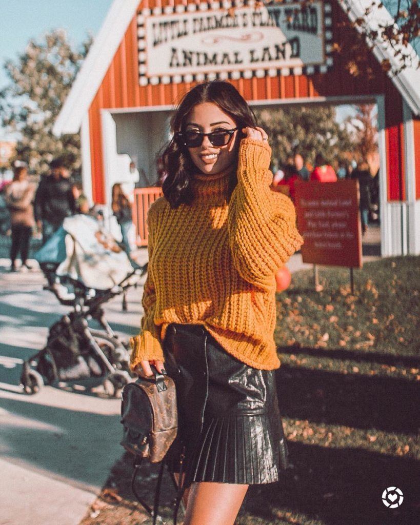 Mulher de cabelo castanho no ombro com óculos de sol, blusa de lã mostarda e saia de couro preta