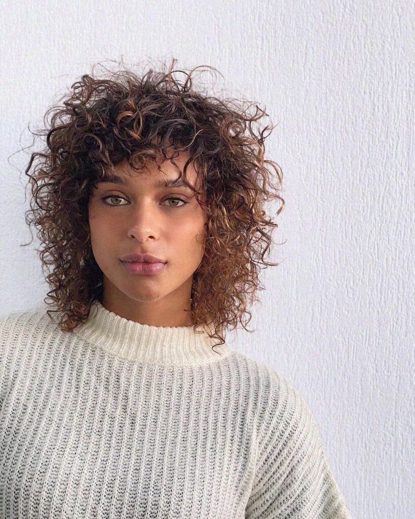 mulher com cabelo médio cacheado pigmaleão usa blusa de lã creme