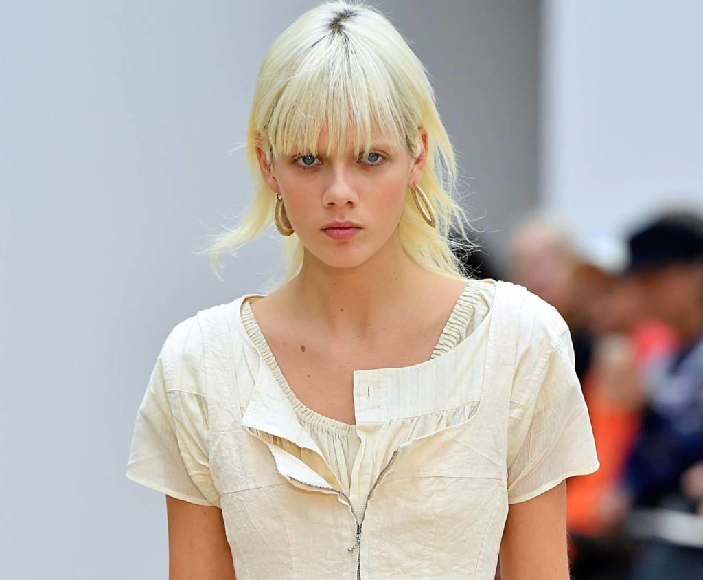 modelo com cabelo platinado e franja desfila com vestido bege e brincos de argola