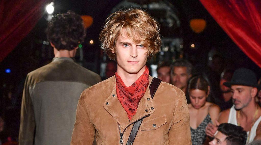 Homem loiro com cabelo liso usa casaco caramelo e blusa vermelha