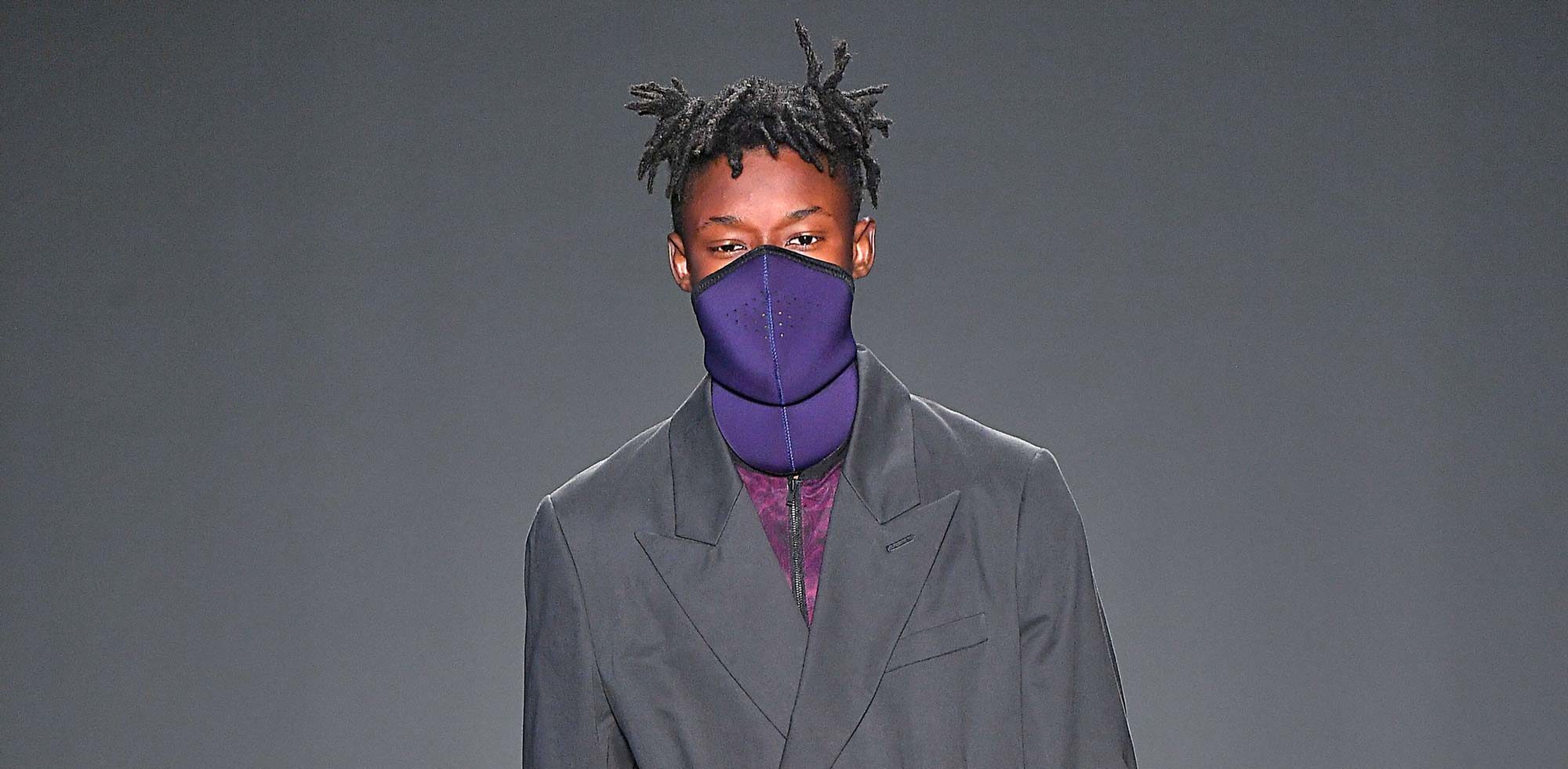 Homem usa dreads no cabelo, enquanto desfila na passarela, usando palitó cinza e blusa roxa.