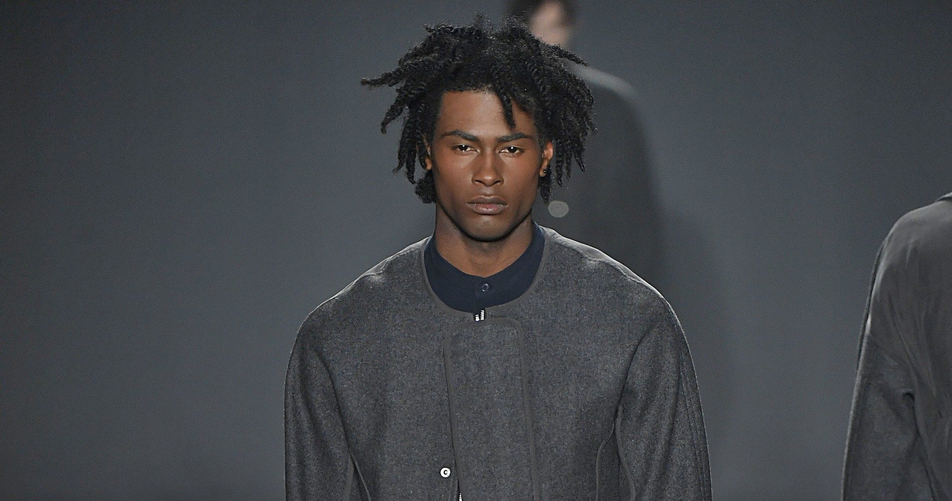Modelo usa cabelo crespo médio e roupa cinza na passarela