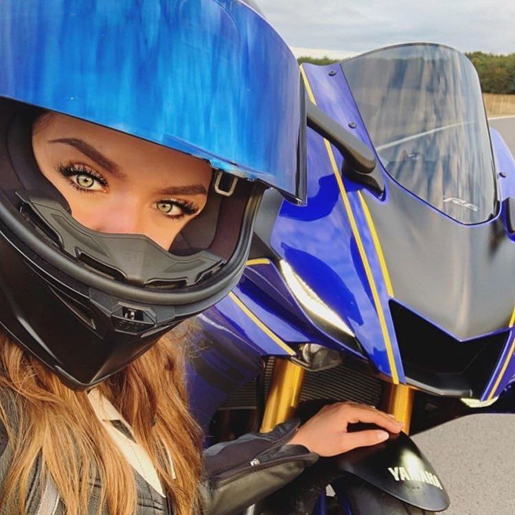 Mulher loira e de cabelos compridos posa com visor do capacete aberto ao lado de uma moto
