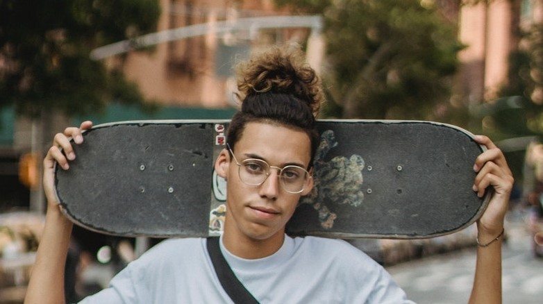 Homem posa com skate e cabelo estilo skatista coque samurai ou man bun no cabelo cacheado castanho