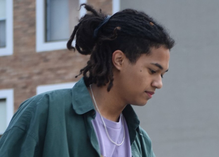 Homem posa ao ar livre com camisa verde e cabelo preto médio com dreads tipo skatista