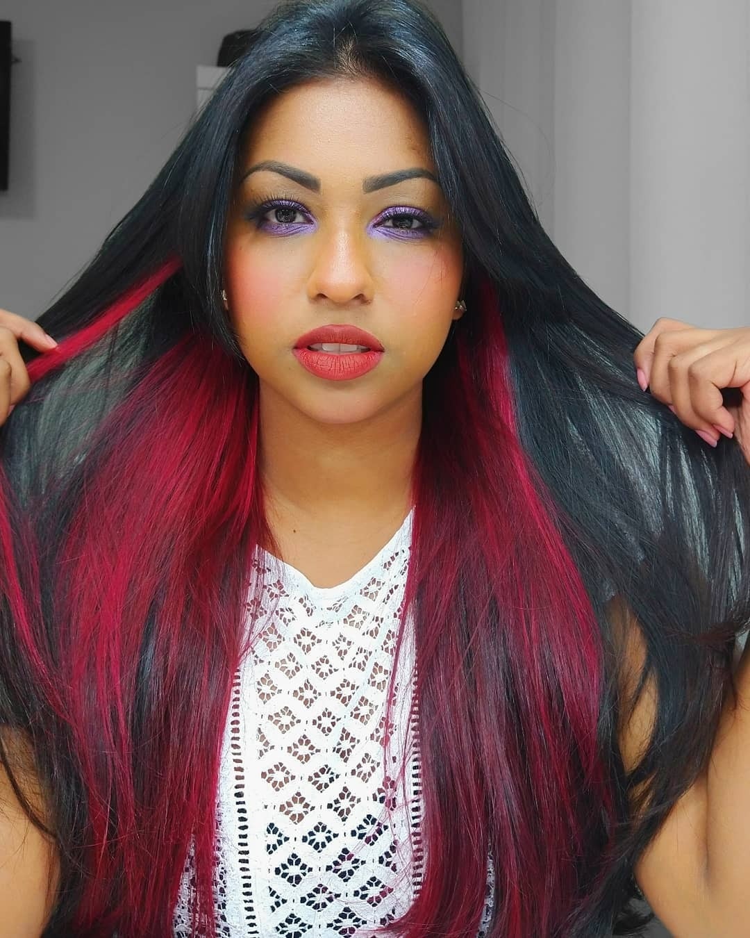 mulherde cabelo preto longo liso posa de frente segurando as pontas dos cabelos com mechas vermelhas em destaque