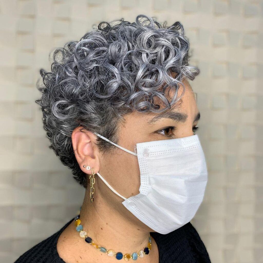 Mulher posa de perfil com máscara e corte de cabelo curto pixie no cabelo cacheado grisalho