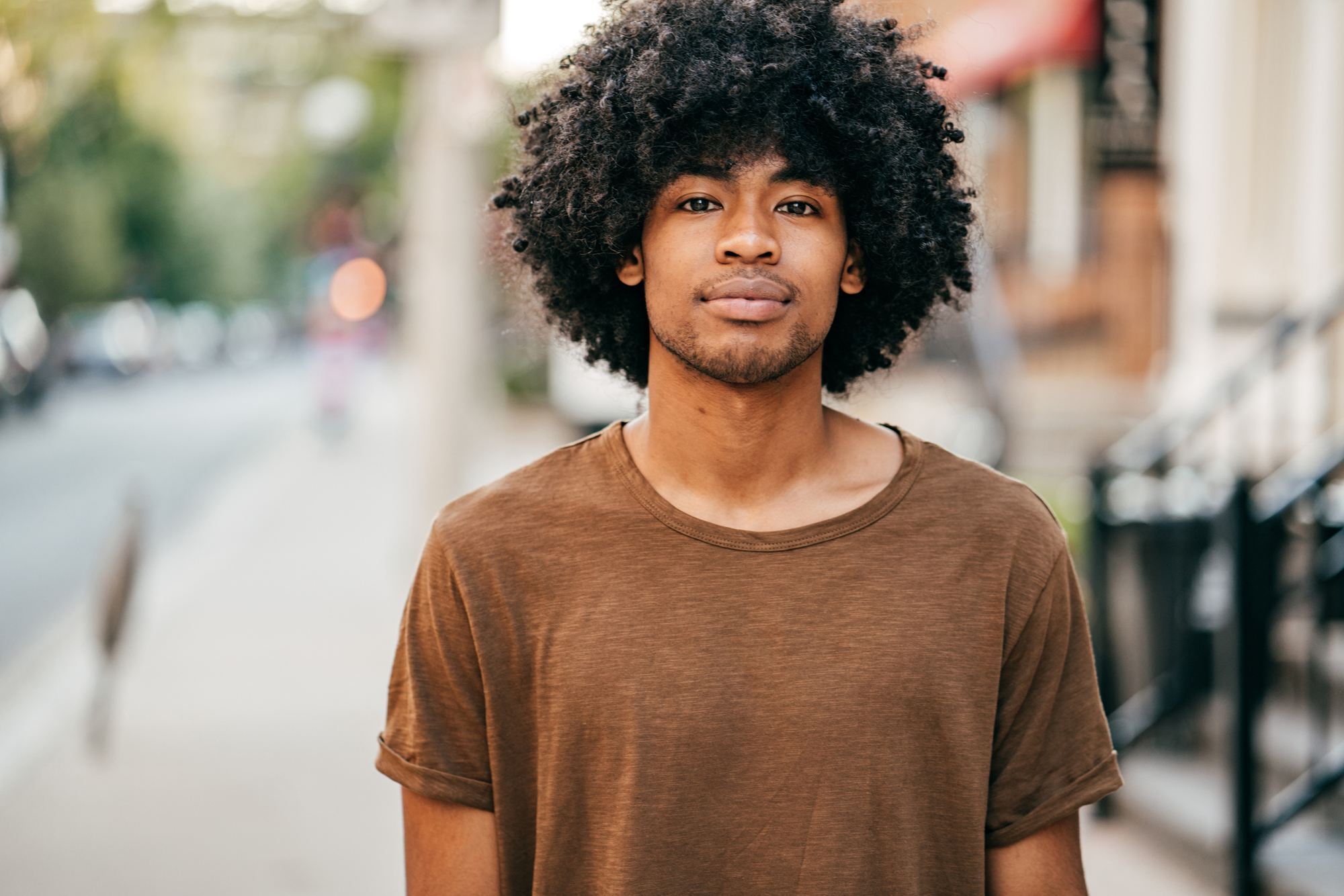 Homem de cabelo volumoso afro cacheado posa ao ar livre de blusa marrom
