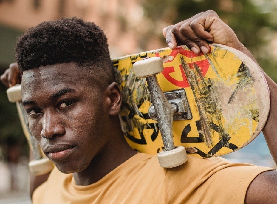 Homem posa de camiseta amarela com skate na mão e cabelo crespo cortado em estilo skatista degradê undercut taper