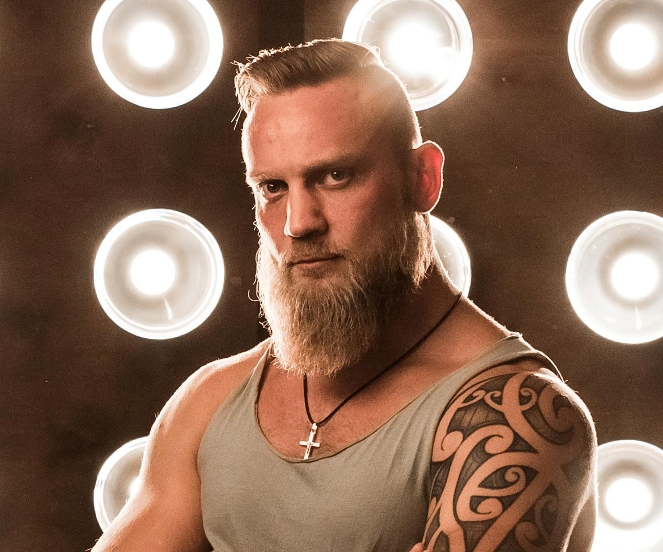 Homem loiro tatuado com barba posa com luzes ao fundo e cabelo undercu