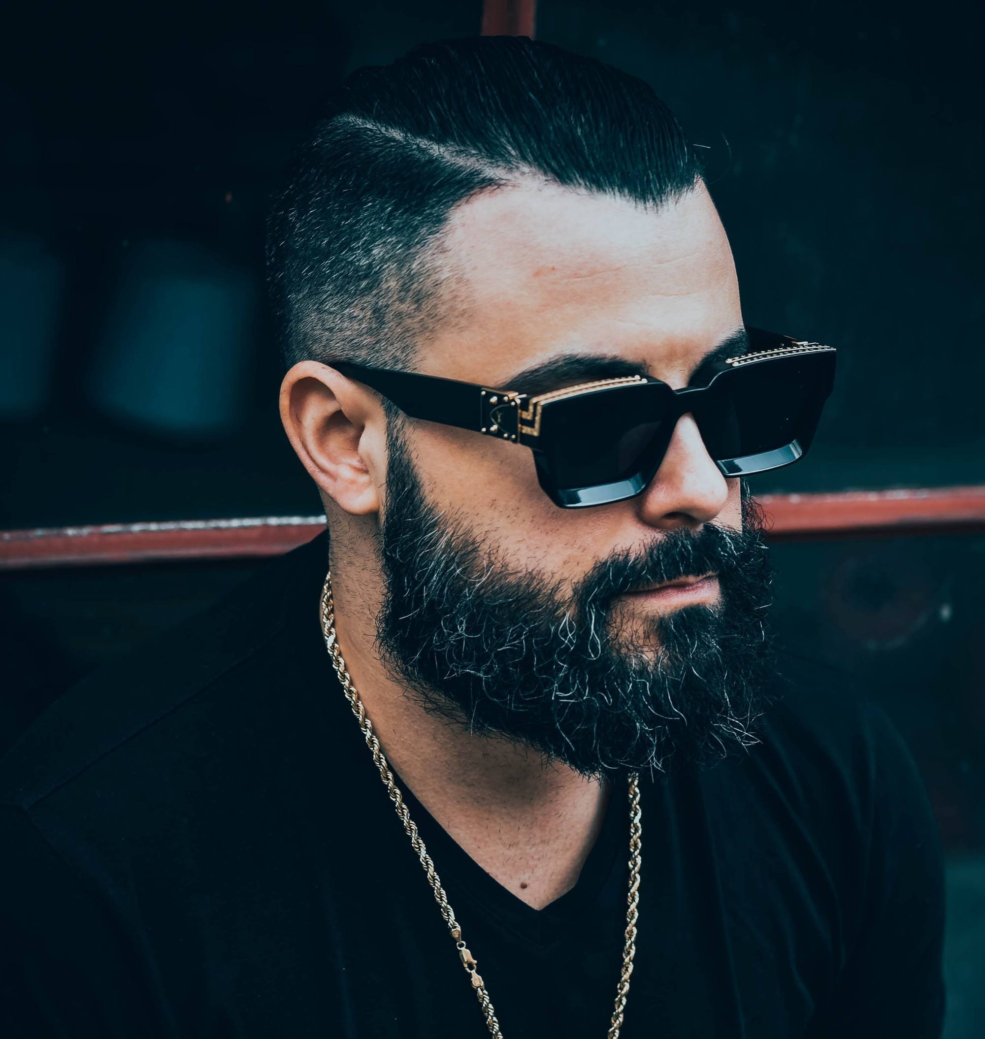 homem usa óculos de sol, corrente e cabelo undercut penteado para trás e barba