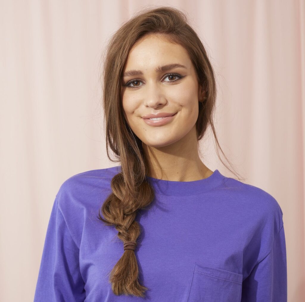 Mulher de blusa roxa posa sorrindo com trança simples lateral frouxa ideal para ir a escola