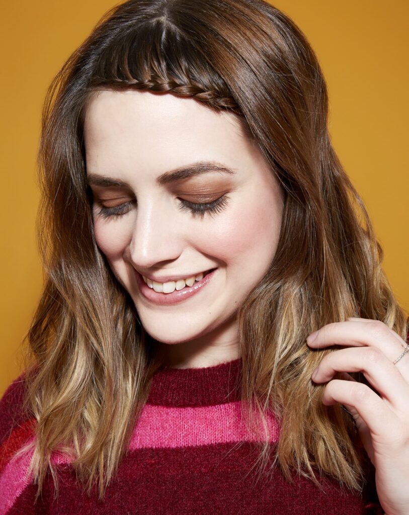 Mulher de blusa vermelha rosa e vermelha posa sorrindo enquanto segura o cabelo com trança simples na franja