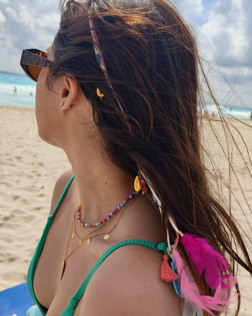 Mulher posa na praia com óculos de sol e tererê com penas cor de rosa na ponta e moedas e pingentes