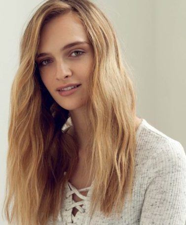 Mulher loira e de cabelos lombos usa blusa branca e cabelo partido de lado e ilustra matéria sobre como riar um look com efeito praia usando chapinha