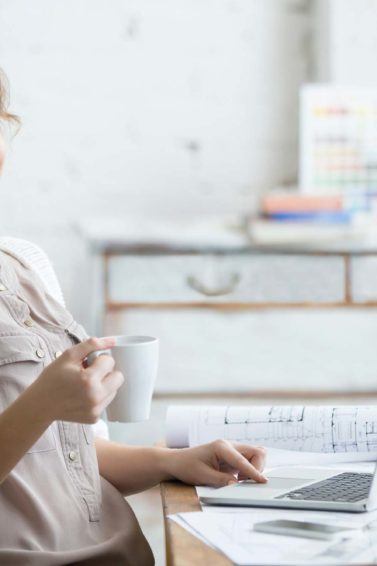 Mulher loira usa roupa clara e toma café em frente ao computador