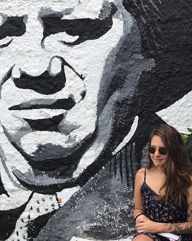 Mulher jovem de cabelos castanhos longos e óculos escuros posa em frente a parede com rosto printado em branco e preto