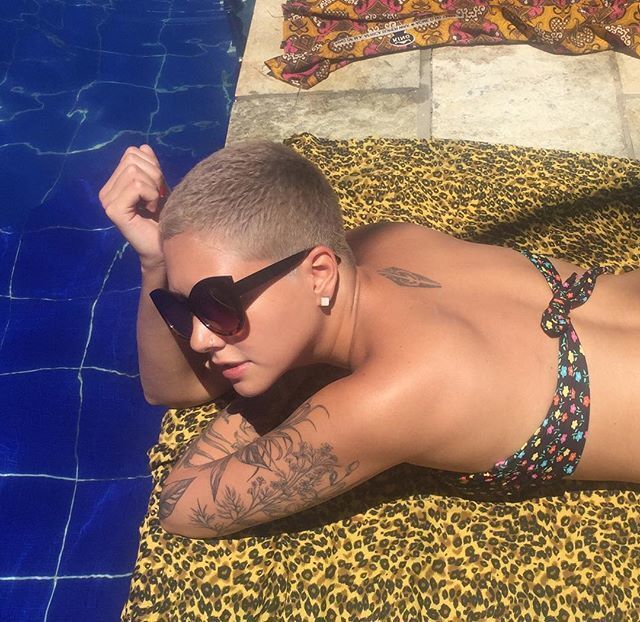 Mulher jovem de cabelo raspado e descolorido toma sol de bruços à beira da piscina sobre canga de animal print