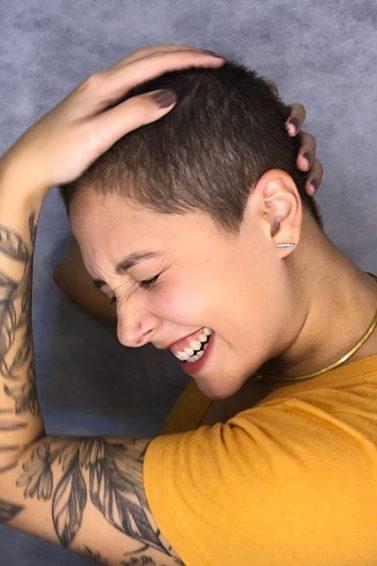 mulher jovem segura a cabeça com cabelos raspados e sorri. Ela tem tatuagem de flores nos braços e camiseta de manhga curta amareça
