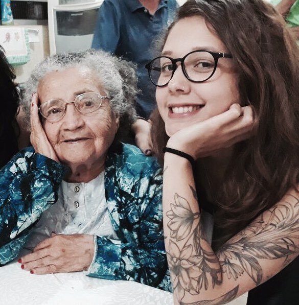 Mulher jovem de cabelos castanhos longos e braço tatuado com flores ao lado de senhora idosa. AMbas sorriem e olham para a câmera. Ela raspou e decidiu doar cabelo