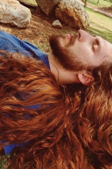 homem de cabelo comprido e acobreado deita na grama de olhos fechados