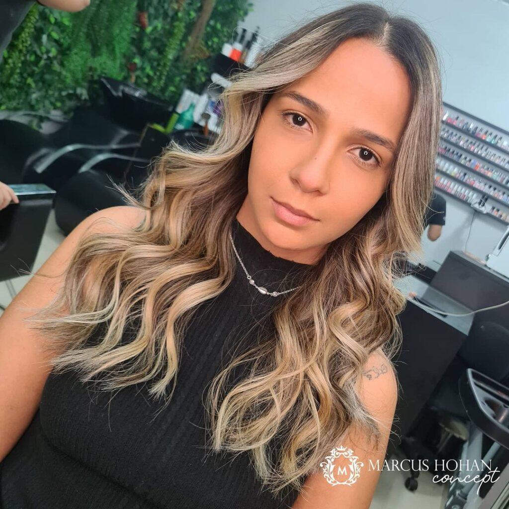 mulher de frente com blusa regata preta e cabelos loiros com raiz esfumada