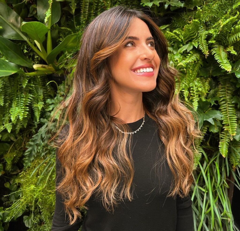 Mulher com blusa preta no salão e cabelo longo em tons de castanho iluminado dourado caramelo e franja cortina