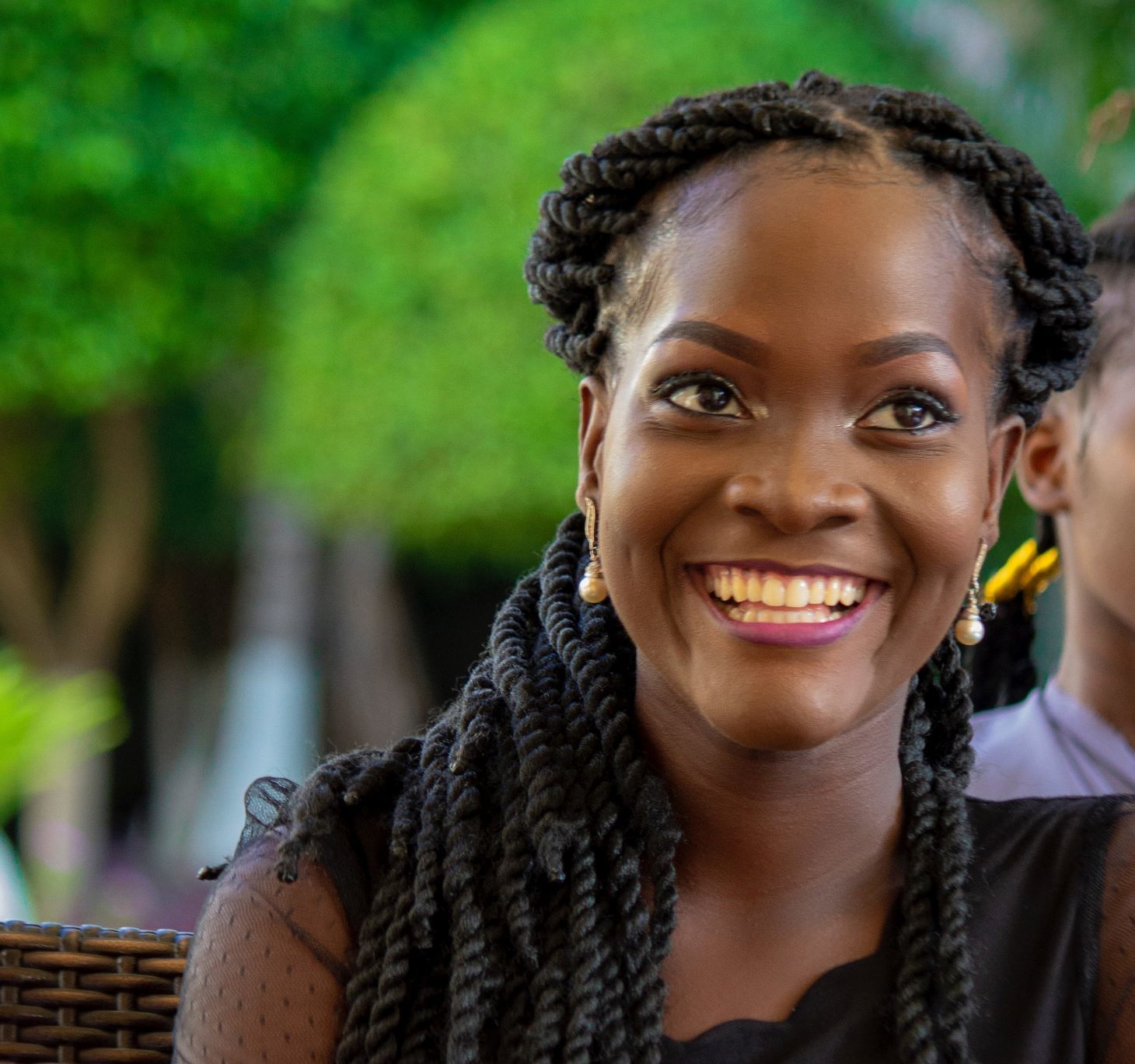 Mulher sorrindo. Ela tem cabelos trançados em um penteado preso para trás e brincos de pérolas