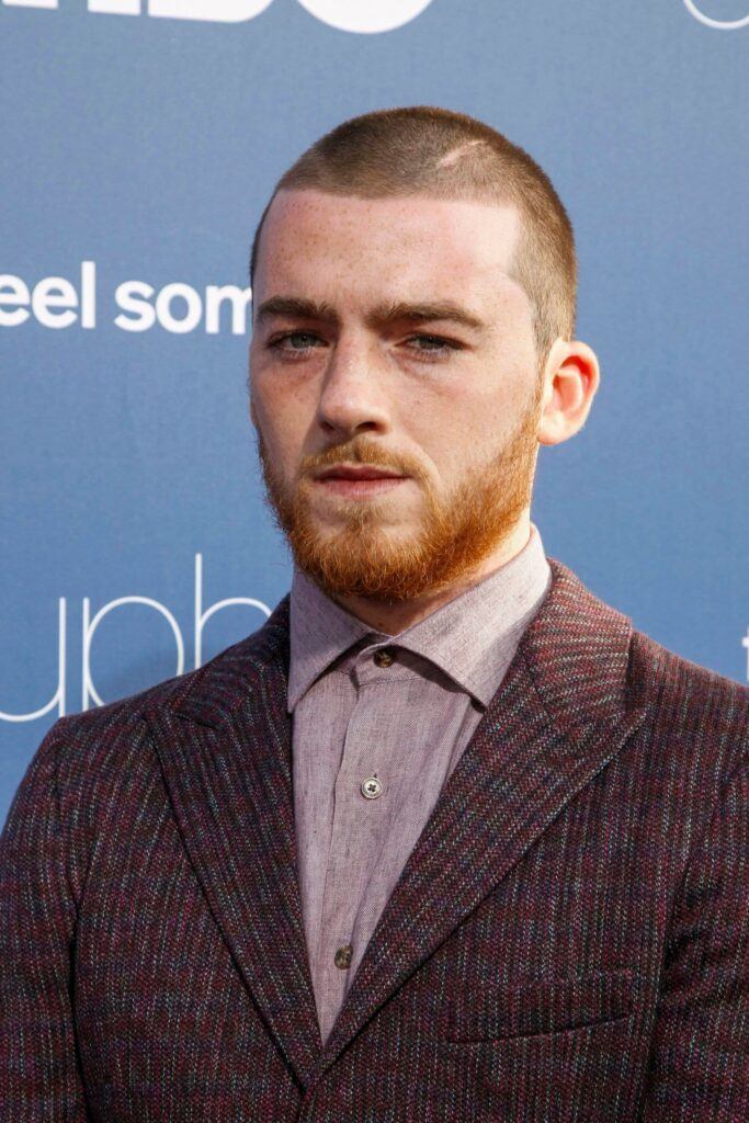Ator Angus Cloud posa em evento de terno vinho e corte buzz cut no cabelo grosso ruivo