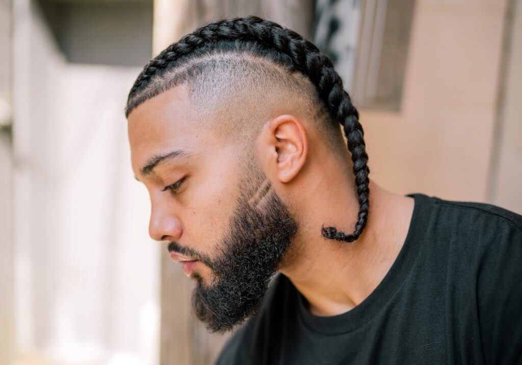 Homem posa de perfil com camiseta preta e tranças undercut no cabelo grosso raspado nas laterais afro cacheado ou crespo