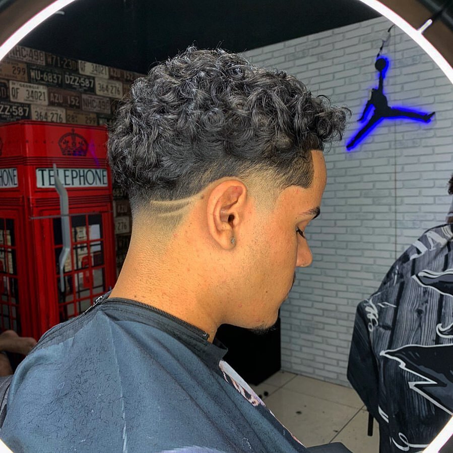 Homem posa de costas no salão com corte taper fade no cabelo grosso castanho ondulado
