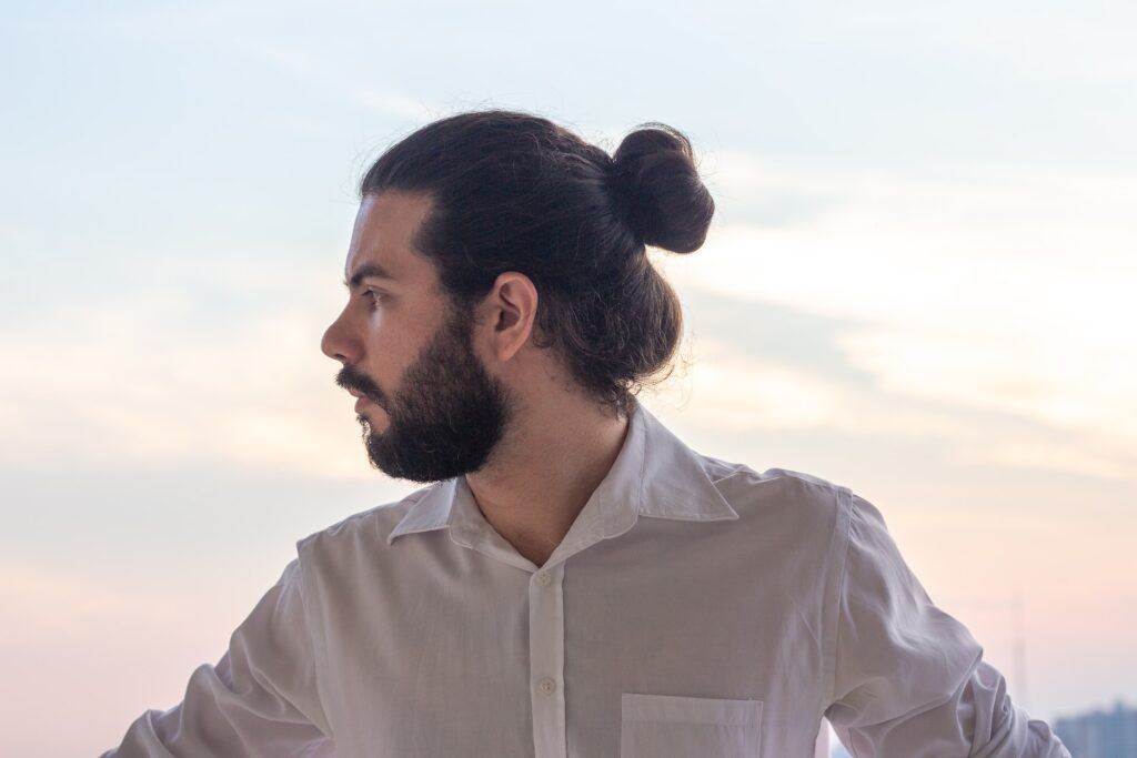 Homem de perfil com camisa branca e coque samurai no cabelo grosso longo liso e castanho