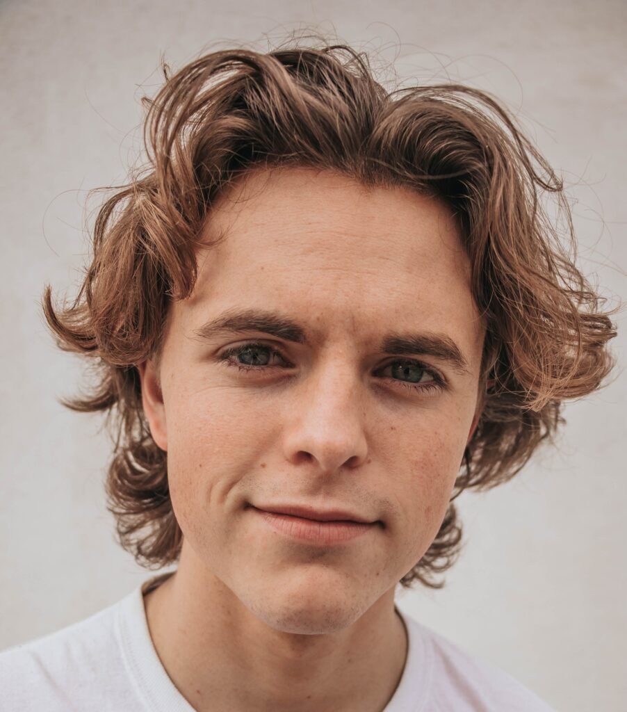 Homem de camiseta branca posa de frente com cabelo grosso loiro solto e ondulado com corte shaggy