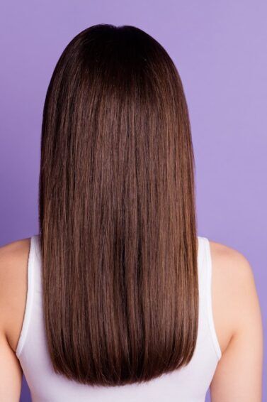 Mulher de costas com blusa branca num fundo roxo e cabelo liso castanho com progressiva sem formol