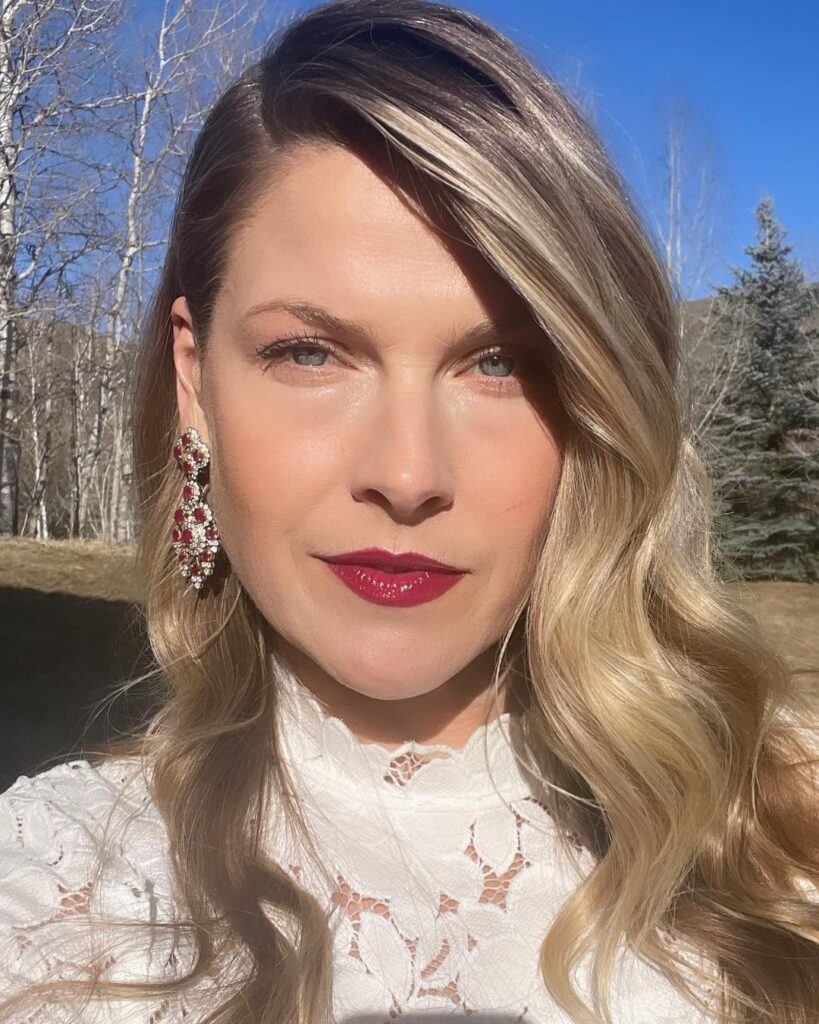 Atriz Ali Larter posa ao ar livre de vestido branco, maquiagem e cabelo loiro ondulado no rosto triangular