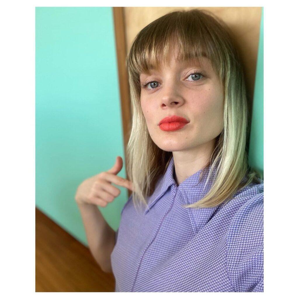 Atriz Bella Heathcote posa com camisa lilás e cabelo long bob com franja loiro com luzes no rosto triangular