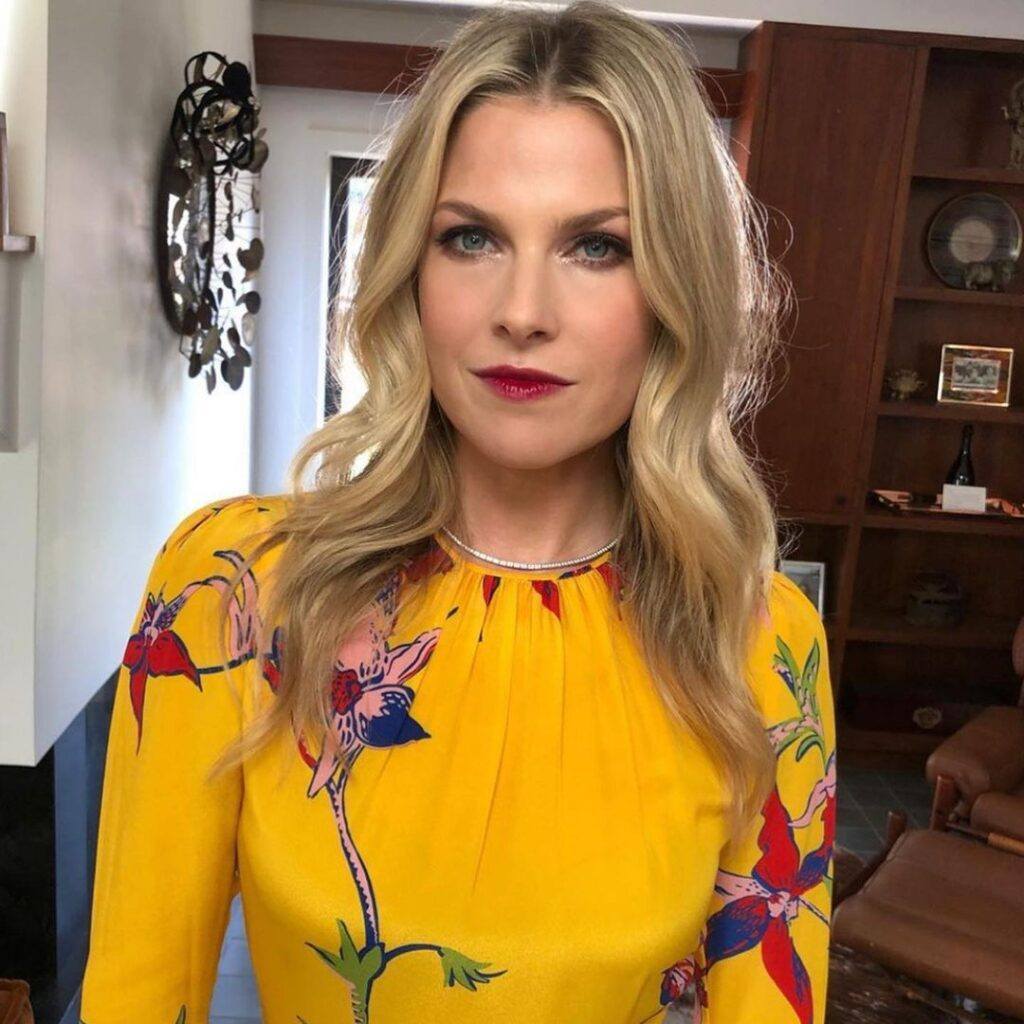 Atriz Ali Larter posa de vestido amarelo florido, maquiagem e cabelo loiro ondulado no rosto triangular