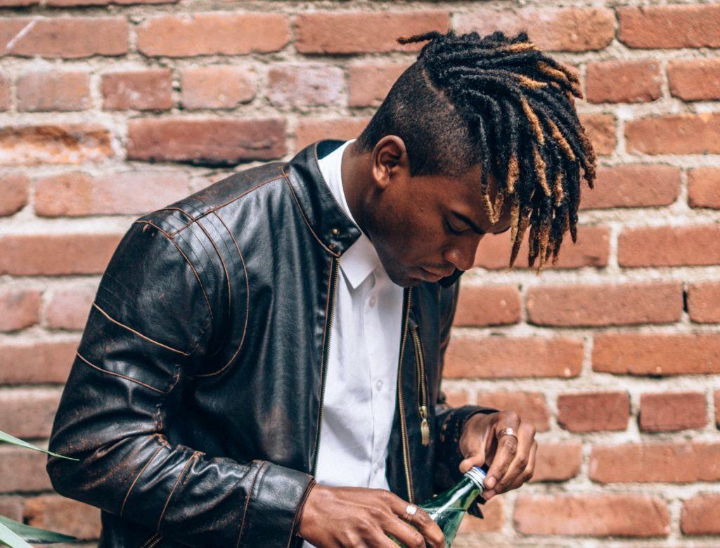 Homem com cabelo com dreads, na rua, em frente a uma parede de tijolos